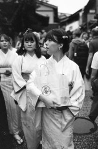 Black and white photo of three women wearing traditional kimonos walking through a bustling street in Kyoto. The woman in the front carries a bag with Japanese writing, and the street is crowded with people in the background.