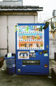 A blue vending machine stands outdoors in an urban area of Kyoto, offering a variety of beverages including tea, coffee, and energy drinks. Adjacent to it is a trash bin with a plastic bag liner. In the background, buildings and rooftops are visible, capturing the essence of Japon in Octobre 2019.