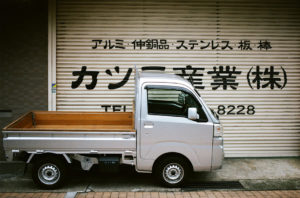 A small silver pickup truck is parked in front of a gray, closed garage door with Japanese text and contact information written on it. The street in Kyoto consists of tan-colored tiles. This scene captures a typical moment from Japon in Octobre 2019.