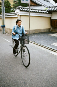 A woman wearing a blue jacket and a backpack rides a bicycle with a front basket on a quiet street in Kyoto. She is passing by traditional Japanese buildings with tiled roofs and beige walls, capturing the serene essence of Japon in Octobre 2019.