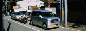 A small, silver car is parked on a quiet street in Takayama alongside a motorcycle and another car. There is a building in the background with a sign and a visible "P" parking sign. The street appears clean, and the weather is sunny, capturing the serene atmosphere of Japon in October 2019.