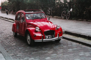 A vintage red 2CV is parked on a cobblestone street next to a pedestrian walkway. In the background, a few bicycles are secured to a metal railing, and some trees are visible, making it the perfect setting for a nostalgic balade.