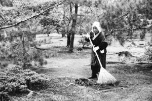 Black and white photo of an elderly woman wearing a headscarf and traditional attire, raking in a serene wooded area. Taken in Kyoto, Japan, October 2019, she is surrounded by trees and appears to be tending to the garden floor covered with leaves and pine needles.