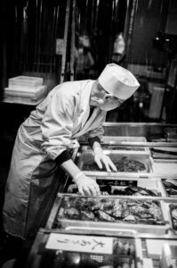 A person in a chef's hat and uniform, wearing gloves, inspects seafood displayed in trays at a market or kitchen. Captured in black and white with a classic, timeless feel, this scene from Kyoto in October 2019 shows a busy, cluttered environment in the background.