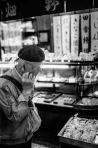 A black and white photo captures an elderly person with glasses and a beret, closely inspecting items on display in a store in Kyoto. The background features a shelf with various goods and signage adorned with Japanese characters, offering a glimpse into daily life in Japon during Octobre 2019.