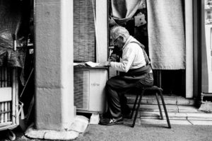 A black and white photo of an elderly man sitting on a small stool outside a shop in Kyoto, focused on writing or sketching on a pad. He wears glasses, suspenders, and is engaged in his work near a Fujitsu air conditioning unit. The shop's door is partly covered with a curtain. October 2019.