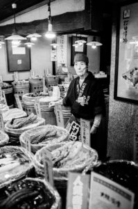 Black and white photo of an elderly vendor wearing traditional attire standing inside a market stall. Tubs of assorted goods, possibly pickles or preserved foods, are displayed in front of the vendor. Japanese signage and framed pictures adorn the walls. Taken in Kyoto, Japon, octobre 2019.