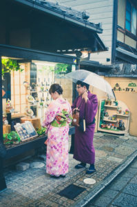 A man and woman, both dressed in traditional Japanese kimonos, stand outside a shop window on a narrow, stone-paved street in Kyoto. The man holds a transparent umbrella. Shelves with various items and plants are visible inside the shop.