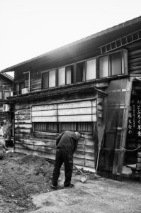 A person wearing dark clothing bends over to sweep in front of a rustic wooden building with several windows and sliding doors. A broom leans against the wall, and Japanese text is visible on a transparent panel beside the entrance. The scene, captured in black and white, evokes the timeless charm of Shirakawago in Japon, Octobre 2019.