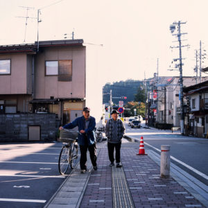 Two elderly men walk along a sidewalk in Takayama, with one pushing a bicycle. They pass buildings and a few parked cars on a sunny October day in 2019. Street signs and utility poles are visible, with a small construction cone at the intersection. The scene has a calm, everyday life atmosphere.