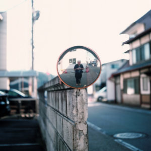In Takayama, Japon, a convex mirror mounted on a concrete wall reflects a person standing on a quiet street lined with buildings. The scene from Octobre 2019 is bright with clear visibility, showing parked cars on one side and houses on the other.