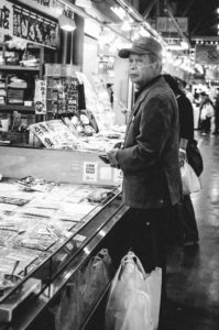 A black-and-white image shows an elderly man wearing a cap and jacket, standing in a store aisle at a Marché aux poissons in Shizuoka, Japon. He looks towards the camera while holding a shopping bag. The aisle is lined with merchandise and lighted from above. Another person is seen in the background.