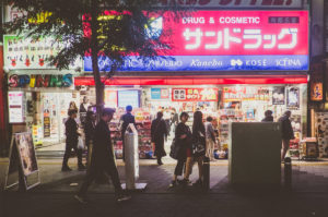 A bustling street scene in Ikebukuro, Tokyo at night, with pedestrians walking past a brightly lit drug and cosmetic store. The store's neon signs and product displays create a vibrant atmosphere. Other shops and advertisements are visible in the background, capturing the essence of Japon nightlife.
