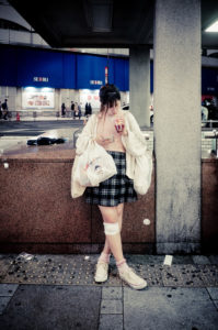 A young person with a casual fashion sense stands at a street corner in Ikebukuro, sipping a drink. They are dressed in a plaid skirt, cozy sweater, and sneakers, with a white knee brace on one leg and carrying bags. The bustling Tokyo city streets with lights and pedestrians form the backdrop.