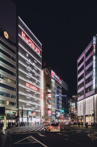 A bustling city street at night in Ikebukuro, Tokyo, illuminated by bright lights from various buildings. A prominent building display reads "BIC CAMERA" in large red letters. Neon signs and billboards in Japanese line the street, with cars and pedestrians moving about.