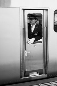 A train conductor, dressed in a formal uniform with a cap and white gloves, stands in the doorway of a sleek, modern train on Tokyo's metro. They appear to be ready for departure or arrival at a station, looking out attentively. The image is in black and white.