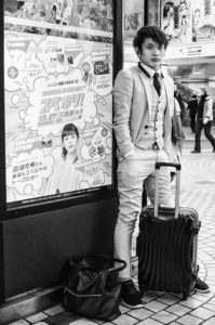 A man in a light-colored suit stands casually against a wall with a large cartoon-themed advertisement in Tokyo, 2019. He is leaning on a black suitcase. The urban setting appears to be a busy area with pedestrians in the background. The scene is in black and white.