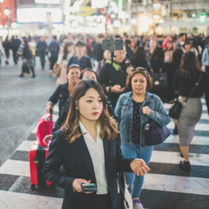 A woman in a suit crosses a busy street with her phone in hand. Several people are walking around her, including someone pulling a red suitcase. The background is bustling with city lights and pedestrian activity.