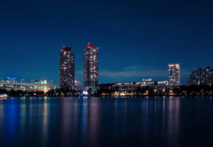 A nighttime cityscape in Tokyo features tall buildings with illuminated windows next to a calm body of water. The lights from the buildings and surrounding structures reflect brightly on the water's surface. The clear sky adds to the serene ambiance, capturing Japan's beauty in October 2019.