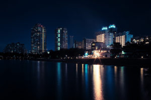 Night view of a Tokyo cityscape with illuminated high-rise buildings reflecting on the calm water in the foreground. In this serene Japon scene from October 2019, the city lights create vibrant streaks of color on the water surface, enhancing the tranquil atmosphere.