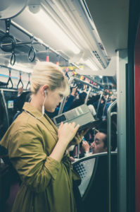 A person with blonde hair, wearing a brown trench coat, stands on a crowded Tokyo subway train reading a book. La femme has earphones in and appears focused on the book. Other passengers are seated and standing around. The overhead straps indicate a busy commute.