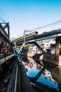 A train travels over a blue elevated bridge above a canal lined with buildings in the Akihabara District on a clear day. The water in the canal reflects the bridge and surrounding structures, creating a serene urban scene in Tokyo.