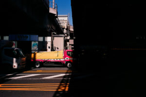 A vibrant pink truck covered with a yellow tarpaulin drives through a city street in the bustling Akihabara District, partially shaded by the overpass above. Another white truck is visible in the brighter area on the left side, and buildings are in the background under Tokyo's clear blue sky.