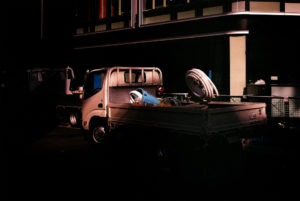 A small white truck is parked at the side of a dark building in Tokyo's Akihabara District. The truck's cargo bed contains coiled hoses and other equipment. Another truck is visible in the background. The scene is dimly lit, with shadows casting across the vehicles and the surrounding area, evoking a 2019 essence.