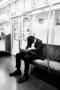 A person in a suit sits on a subway bench with their head resting on a briefcase held on their lap. The Tokyo métro car is empty, and overhead handrails are visible. The person appears to be asleep or resting. The scene is in black and white, capturing the serene solitude of Japon.