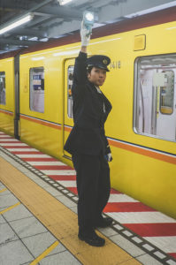 A uniformed train conductor stands on a Tokyo Métro platform beside a yellow train in Japon. The conductor is holding up a signaling device and looking forward. The platform features distinct red, white, and yellow markings.