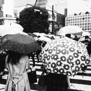 Black and white photo of a busy crosswalk in Shibuya, crowded with people holding umbrellas. A variety of umbrella patterns are visible, including polka dots and flowers. Buildings and trees are seen in the background, and the wet ground suggests it’s raining in this bustling Tokyo district.