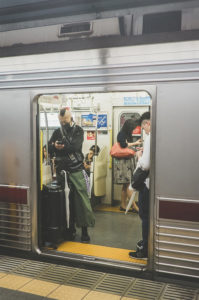 A person with a mohawk hairstyle stands at a Tokyo métro train door, looking at their phone with a suitcase beside them. Other passengers can be seen inside the train, and another person on the platform is also looking at their phone. The train is at a busy Japon station platform.