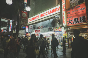 A bustling nighttime street scene in Ikebukuro, Tokyo, filled with numerous people walking past brightly lit storefronts. A prominent sign reads "BIC CAMERA," indicating an electronics store. The atmosphere is lively, with various neon signs and advertisements illuminating the area.