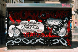 A graffiti-covered metal shutter in Gràcia, Barcelone, displays vibrant artwork and text. The piece features "KIOSKOKUPAT" in large red letters, with additional phrases and graphics including chains, a fist, and eggs. The scene is set in an urban environment.