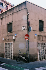 A corner building with beige walls and two graffiti-covered, grated doorways stands in the heart of Gràcia, Barcelone. The structure features various signs, including street names and traffic signs. A bicycle is parked against the wall near a green bike lane on the sidewalk.