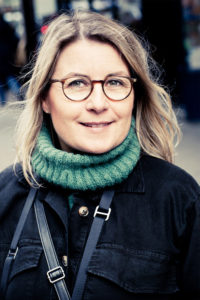 A woman with shoulder-length blonde hair and round glasses smiles while standing outdoors in a green knitted turtleneck sweater. Photographed by Emmanuelle Radenac, she wears a dark jacket with an out-of-focus urban rue backdrop.