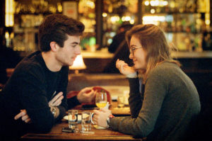 A young man and woman are sitting across from each other at a dimly lit bar, engaged in a conversation filled with charm. They are smiling and gazing warmly into each other's eyes, the air tinged with subtle séduction. The table between them holds drinks and glasses of water, adding to the intimate atmosphere.