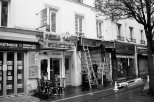 A black and white photo of a street scene in front of a pizza restaurant. Two workers on échelles appear to be installing a sign above the restaurant. One storefront next to it is closed with a metal shutter, and there are several chairs and tables outside the pizza place.