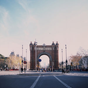 A large, ornate archway structure stands at the end of a broad boulevard in Barcelone, bustling with pedestrians and cyclists. The clear blue sky overhead is dotted with light wisps of clouds, and trees with sparse foliage line the avenue leading up to the arch, reminiscent of picturesque El Born even in hiver.