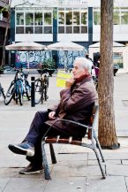 An elderly man sits on a park bench in the Barri Gòtic, resting his cheek on his hand, appearing to be deep in thought. He wears a brown jacket and black pants. Bicycles are parked nearby, and there is graffiti on a wall in the background, capturing the charm of Barcelona even in hiver.