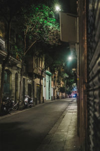 A narrow, dimly lit street in Gràcia at night, lined with parked motorbikes and scooters on the left. Tall buildings with balconies and greenery are on both sides. Warm light glows from some windows and street lamps, casting shadows on the pavement. The winter air keeps the Barcelone street quiet and empty.