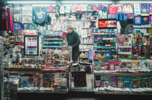 A man stands inside a well-lit newsstand in Gràcia, filled with various items for sale. Shelves are stocked with magazines, drinks, souvenirs, and sports memorabilia. The exterior is decorated with clothing and sports merchandise, including jerseys and hats.