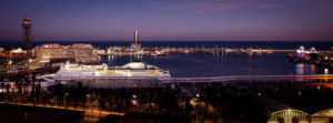 A nighttime view of a busy harbor in Port Vell, Barcelone, lit up with artificial lights. The scene includes a large ferry docked at a terminal, various lit buildings, and docked boats. The horizon shows the dark sea against a twilight sky with hints of pink and blue.