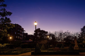 A tranquil park scene at dusk, capturing the essence of heure bleue, with a lit streetlamp in the foreground surrounded by neatly trimmed shrubs. In the background, there's a statue and silhouetted trees against a deep blue and purple sky near Montjuïc, Barcelone. A building with lights is partially visible on the left.