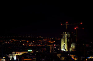 A nighttime aerial view of a cityscape in Barcelone featuring a brightly illuminated Sagrada Familia basilica in the foreground. Two construction cranes stand next to the basilica, with city lights twinkling in the background and a dark sky overhead.