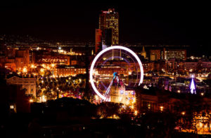 A vibrant night cityscape of Barcelone featuring a large illuminated Ferris wheel at the center. The skyline, typical of a nuit d'hiver, is dotted with various buildings and city lights, giving a bustling and lively ambiance. The background shows a mix of tall, lit-up skyscrapers and a dark, hilly silhouette.