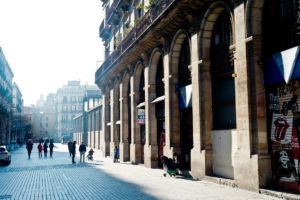A sunlit street in Barcelone's El Born district features people walking and pushing strollers, flanked by a building with tall stone arches on the right. Fachades of the buildings on the left are shaded. The street ends with more buildings in the background under a clear sky.