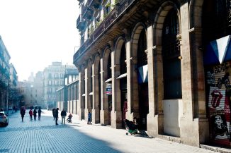 A sunlit street in Barcelone's El Born district features people walking and pushing strollers, flanked by a building with tall stone arches on the right. Fachades of the buildings on the left are shaded. The street ends with more buildings in the background under a clear sky.