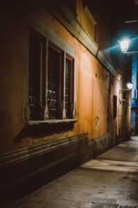 Dimly-lit narrow alley at night with old, weathered buildings on both sides. The left building has barred windows and a worn orange facade. Two street lamps cast a soft blue glow, illuminating the cobblestone path. The alley leads to a brightly lit area, evoking the quiet charm of Barcelone in hiver.