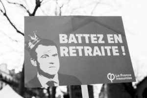 A black-and-white image shows a protest sign featuring a stylized portrait of a man with a drawn-on crown. The sign reads "BATTEZ EN RETRAITE !" and is branded with "La France insoumise" logo, suggesting it is related to the 11 Janvier 2020 manifestation against the réforme des retraites.